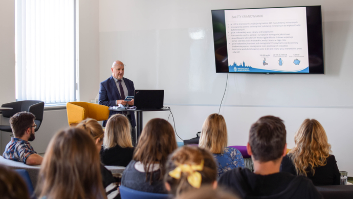 People sit with their backs to the lens on chairs and watch the presentation projected in front of them , which is described by a man in a suit who stands on the left side of the photo